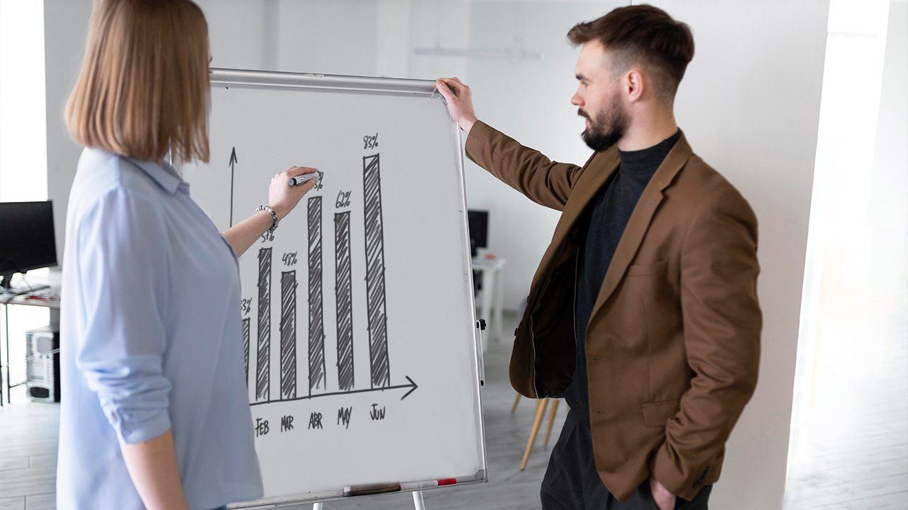 A woman in a light blue blouse and a man in a brown blazer discuss a bar graph drawn on a whiteboard in an office setting. The graph represents data from February to June, showing an upward trend with percentages labeled on each bar. The woman is holding a marker and pointing at one of the bars, while the man holds the whiteboard and listens attentively. In the background, there are office desks, chairs, and computer monitors.