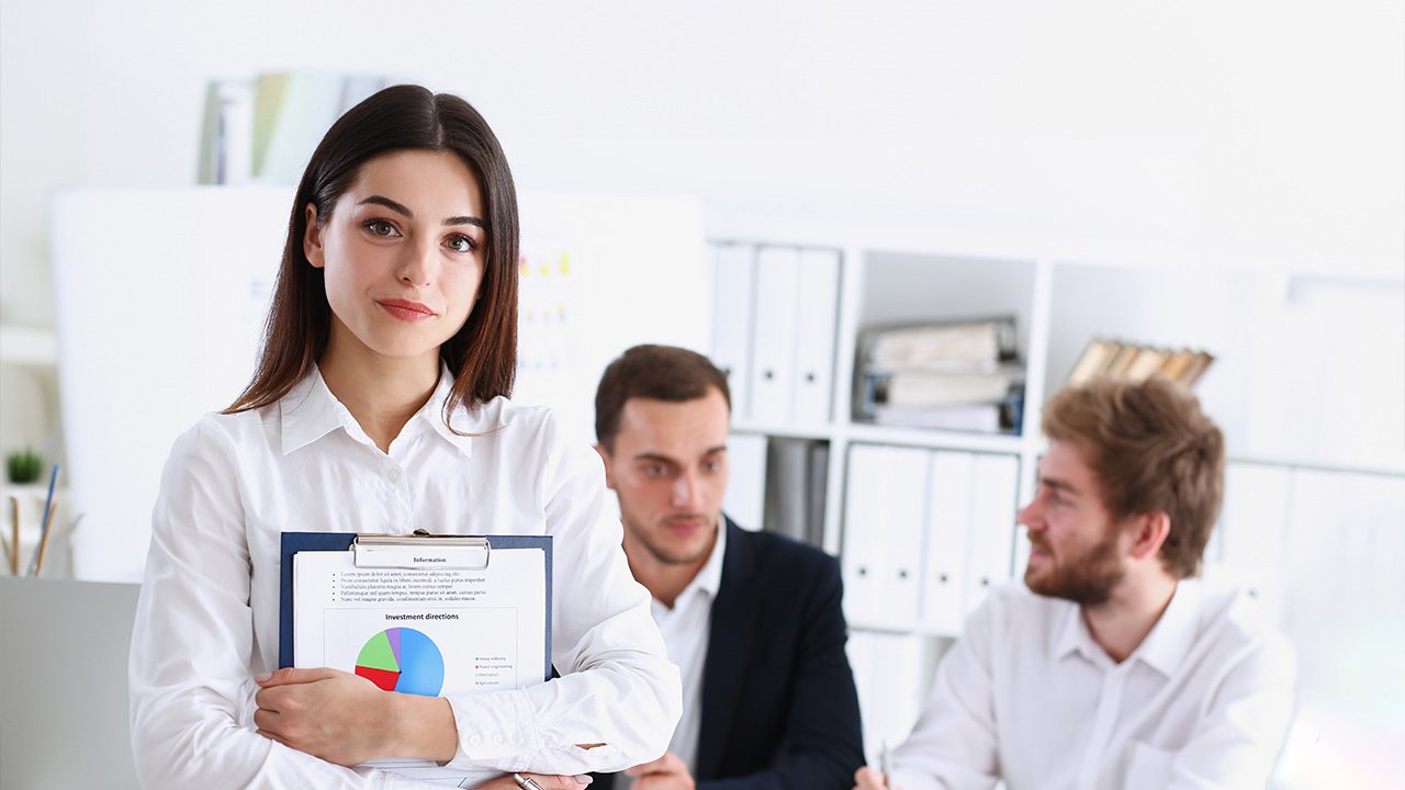 A confident young woman with long dark hair, wearing a white blouse, stands in the foreground of a bright office holding a clipboard with financial charts and documents. Behind her, two male colleagues in business attire are engaged in conversation, slightly out of focus. The office features white shelves filled with binders and books, creating a professional and organized workspace.