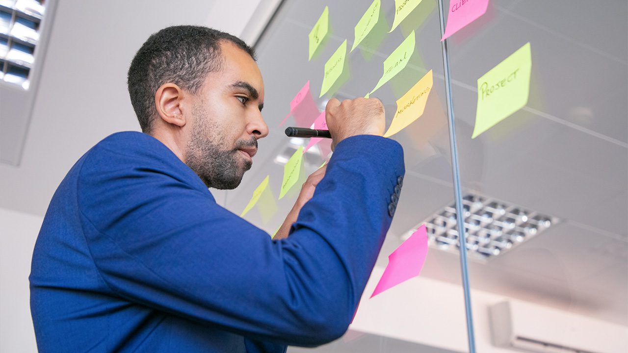 A focused businessman in a blue suit writes on a sticky note with a black marker, adding it to a glass wall covered in colorful sticky notes. The notes contain words like "Project" and "Marketing," suggesting a brainstorming or planning session. The modern office setting features a reflective ceiling and bright lighting, emphasizing a collaborative and strategic work environment.