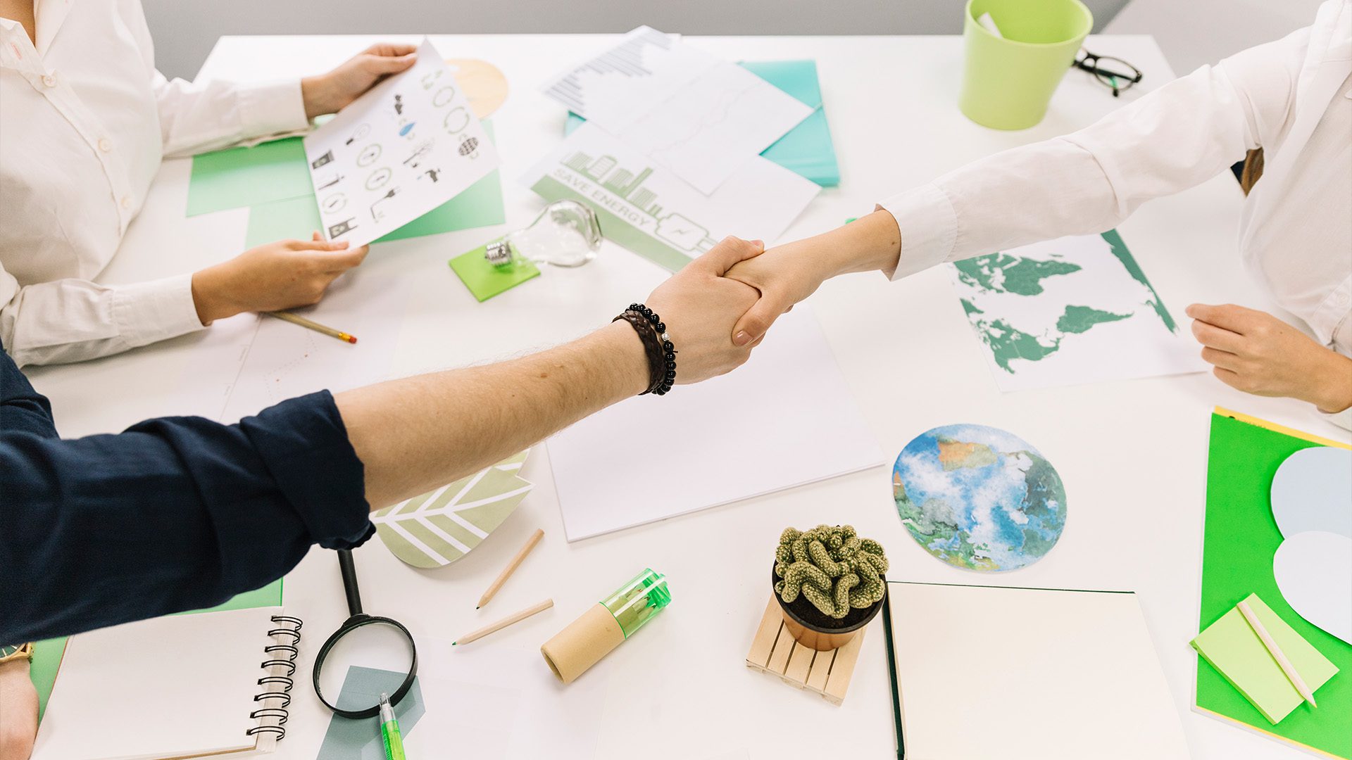 Two people shaking hands over a desk with sustainability-related documents, green-themed office supplies, and eco-friendly materials, symbolizing an environmental agreement or collaboration.