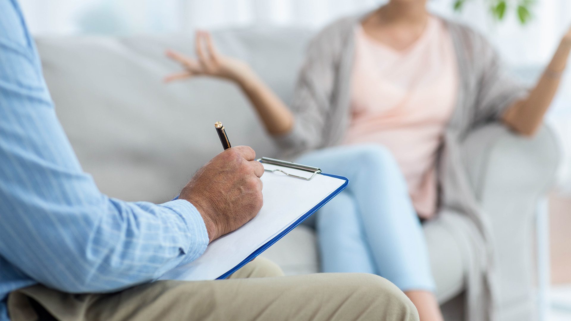 A therapist taking notes on a clipboard while a patient sits on a couch, gesturing with her hands during a counseling session. The focus is on the therapist's hands and clipboard, while the patient is slightly blurred in the background.