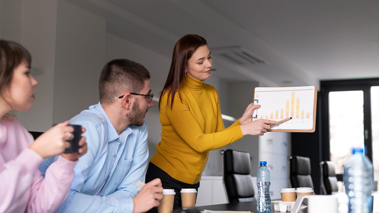 A woman in a mustard yellow turtleneck stands in a modern office, presenting a clipboard with a bar chart to two colleagues. She points at the chart with a pen while the colleagues, a man in a light blue shirt and glasses and a woman in a pink sweater, listen attentively. The table is filled with coffee cups, water bottles, and documents. The office has a contemporary design with black chairs and large windows allowing natural light to enter.
