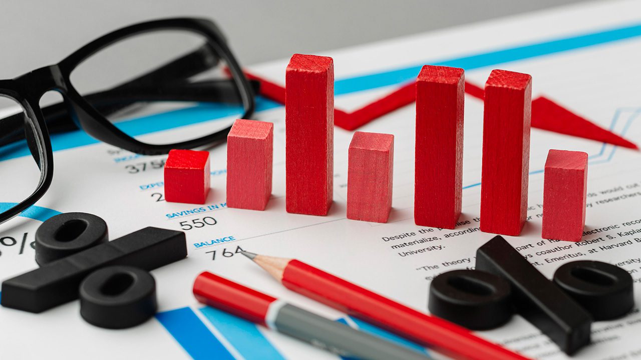 A close-up of financial documents with a miniature red bar chart made of wooden blocks, symbolizing data analysis or business growth. Black percentage symbols, a pair of black eyeglasses, and red and gray pencils are also scattered on the papers. The background includes financial figures and graphs, emphasizing an economic or financial theme.