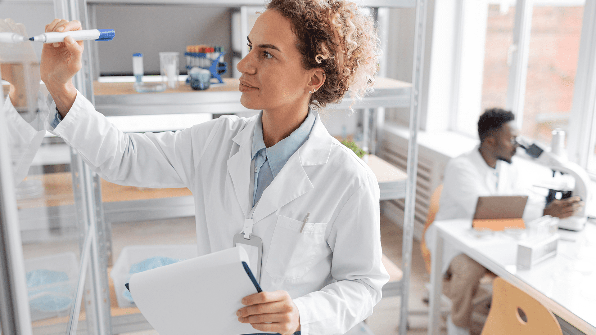 A female healthcare professional in a white lab coat writing on a whiteboard with a blue marker, while a colleague in the background works with a microscope and a laptop on a desk.