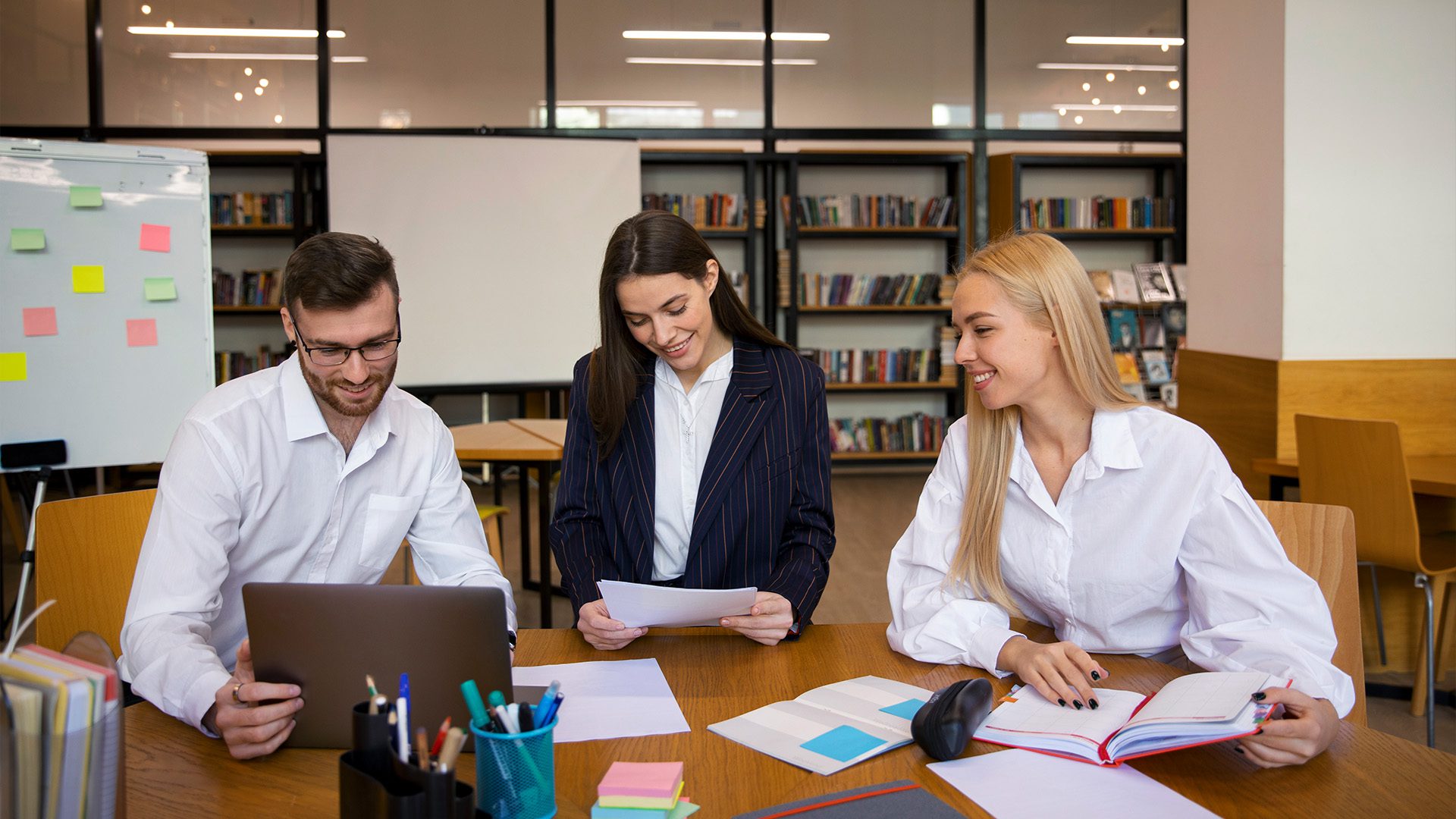 The image shows three professionals collaborating in a modern office or study environment. They appear engaged and happy, suggesting a productive and positive work atmosphere. The setting includes bookshelves, a whiteboard with sticky notes, and office supplies, indicating a brainstorming or team discussion session.