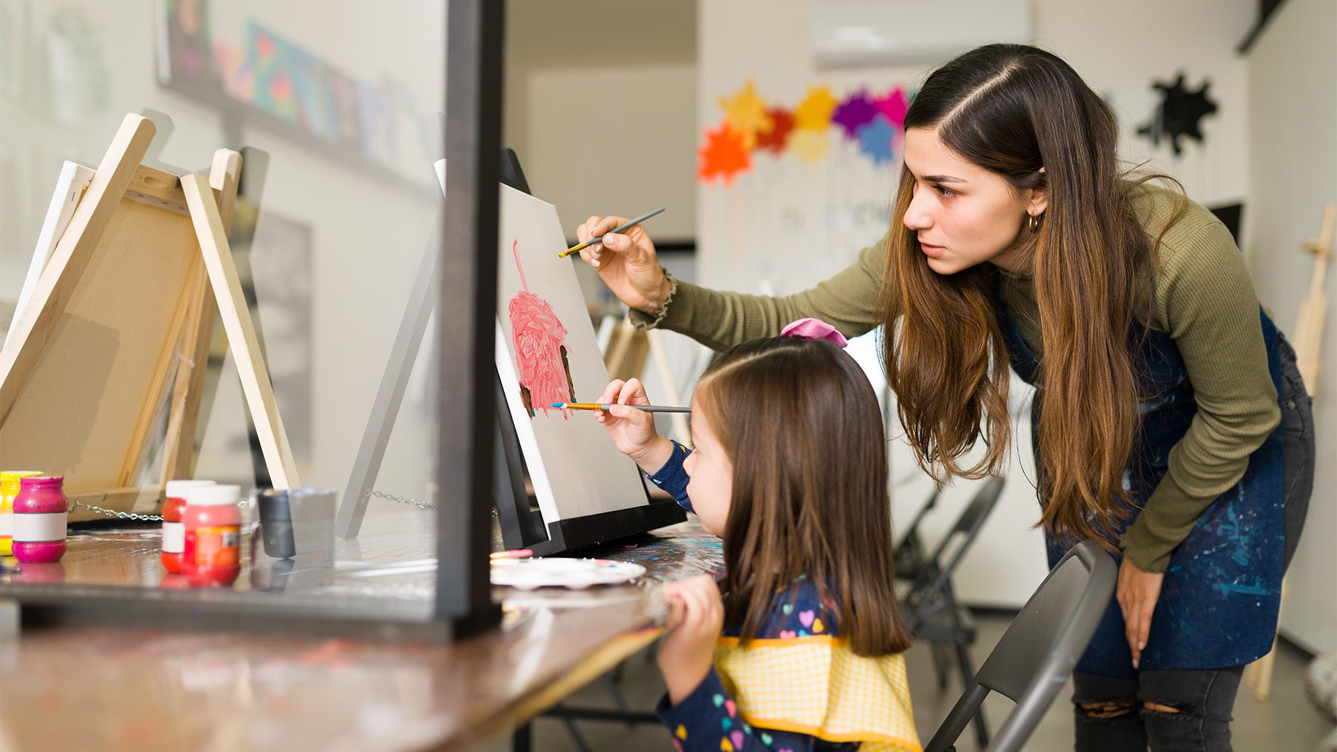 The image shows a young girl painting on a canvas with the guidance of an adult woman, likely an art teacher or a mentor. The setting appears to be an art classroom or workshop, with multiple easels, paint containers, and other art supplies visible on the table. The teacher is leaning in and attentively helping the child with her artwork, suggesting a nurturing and educational environment.