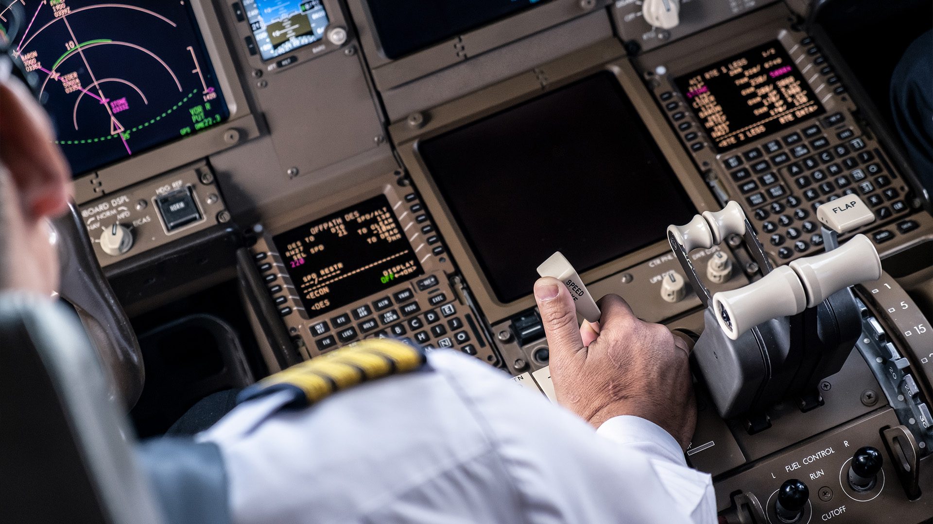 Pilot in uniform operating the throttle controls in an aircraft cockpit, with illuminated flight instruments and navigation screens in the background