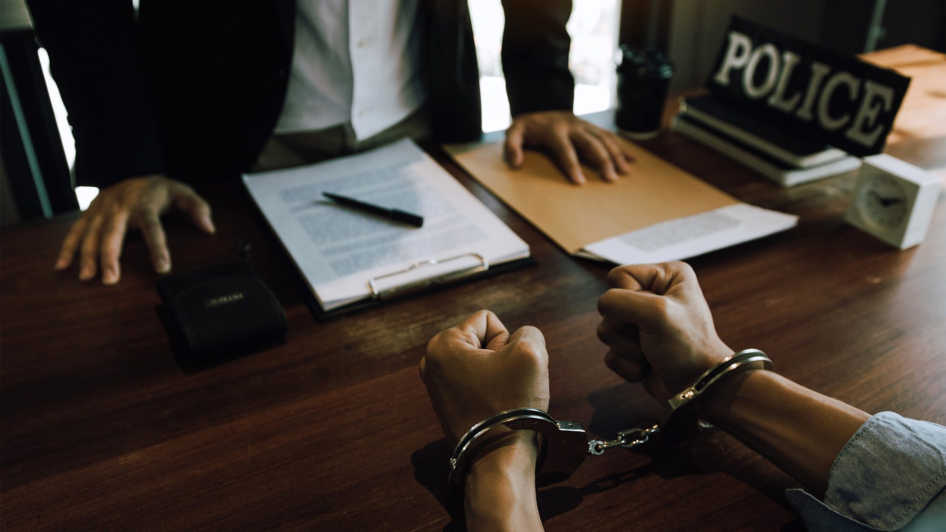 The image appears to depict a scene in a police interrogation or investigation room. A person wearing handcuffs is sitting across from two individuals, likely law enforcement officers or investigators. The setting includes documents, a pen, a folder, and a sign that says "POLICE," indicating an official legal proceeding or questioning.