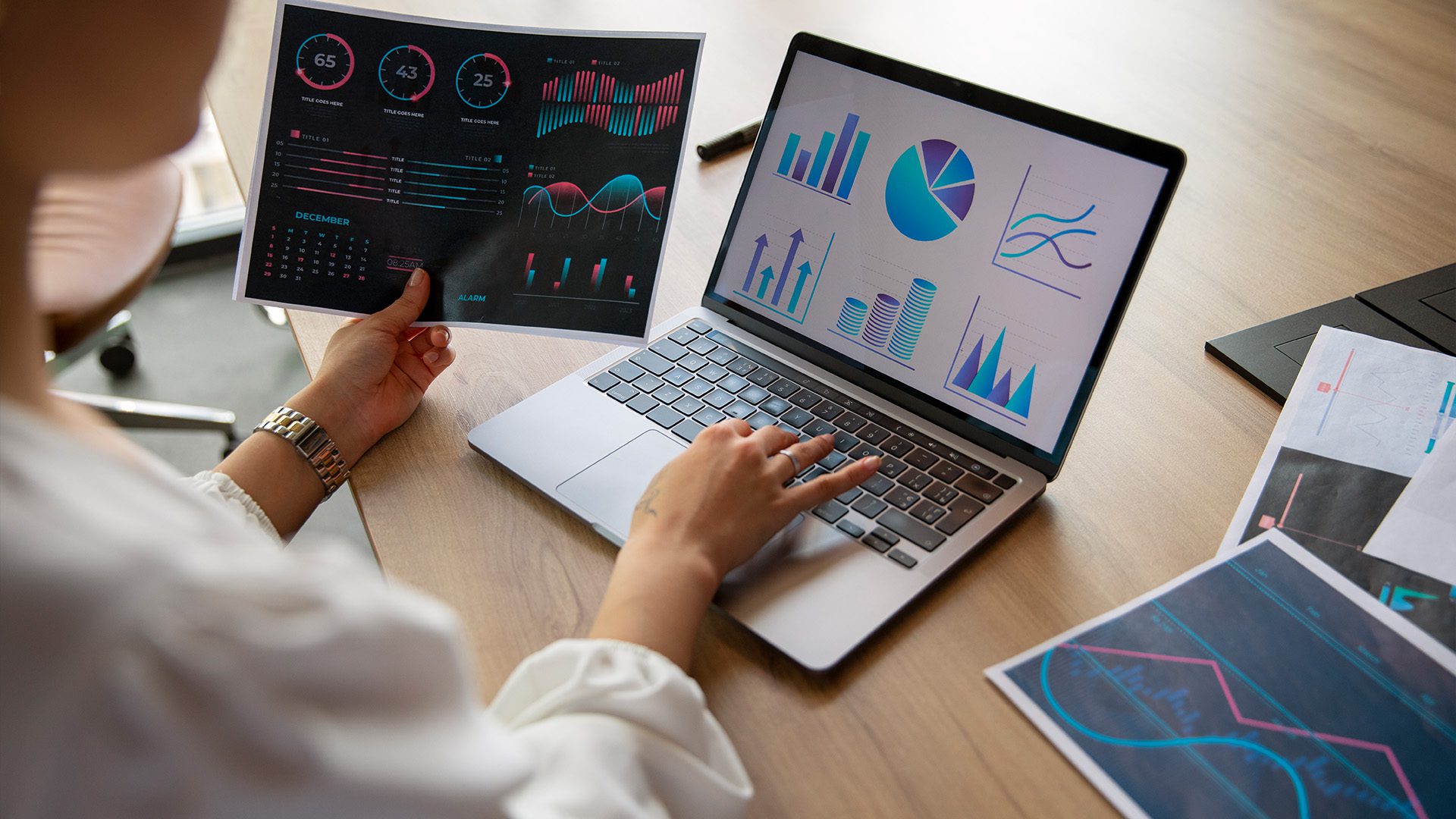 The image shows a person analyzing data visualizations on both a laptop screen and printed documents. The graphics include various charts, such as bar graphs, pie charts, and line graphs, which indicate a data-driven or analytical work environment.