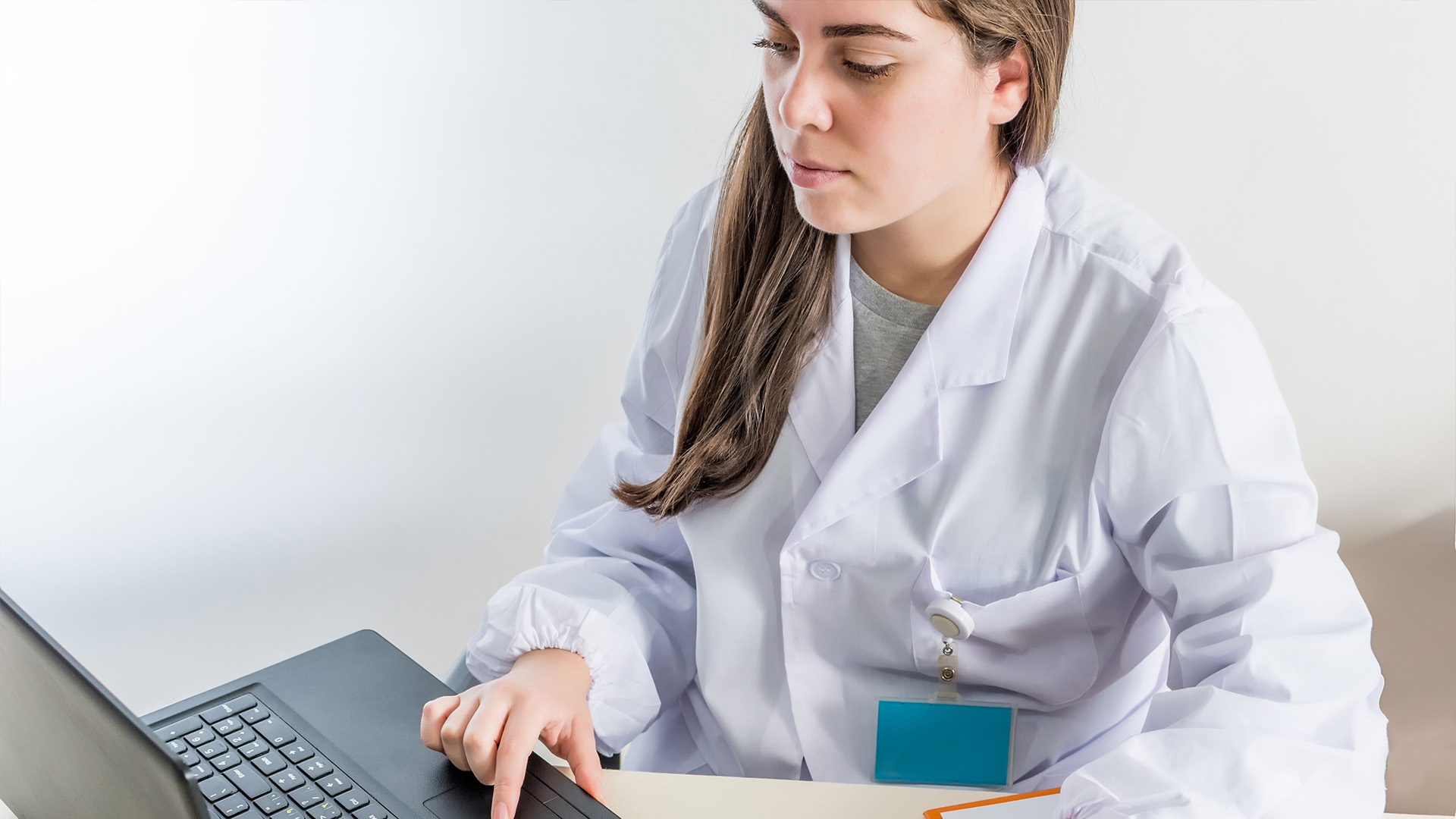 The image shows a woman wearing a white lab coat working on a laptop. She appears focused, possibly engaged in research, data analysis, or medical documentation. The setting suggests a professional or academic environment, likely in a healthcare or scientific field.