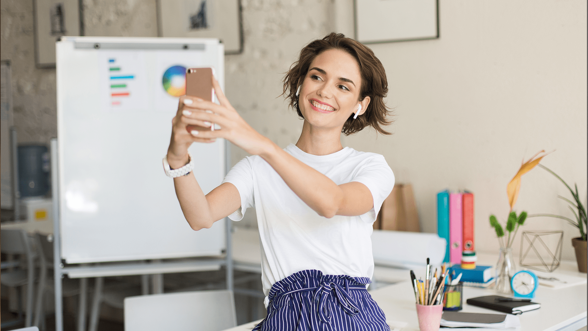 A young woman with short brown hair is smiling and taking a selfie with her smartphone while wearing wireless earbuds. She is dressed in a white t-shirt and high-waisted blue striped pants. Behind her is a bright, modern office space with a whiteboard displaying colorful charts, a desk cluttered with stationery, books, and a small potted plant.