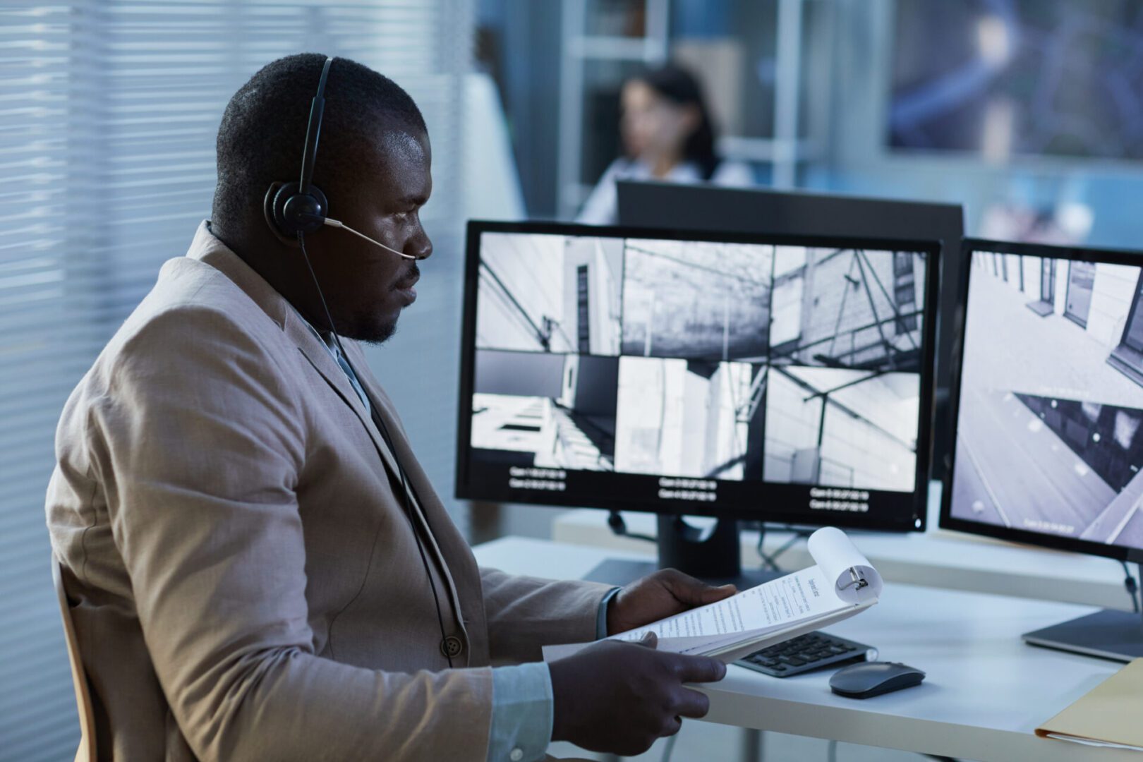 The image shows a man wearing a headset, sitting at a desk in what appears to be a security control room. He is monitoring multiple surveillance camera feeds displayed on two computer monitors in front of him. The feeds show different views of a building or facility. The man is holding a clipboard with documents, possibly taking notes or reviewing incident reports. The setting suggests he is a security officer or surveillance analyst, actively monitoring for unusual activity or managing site security operations.