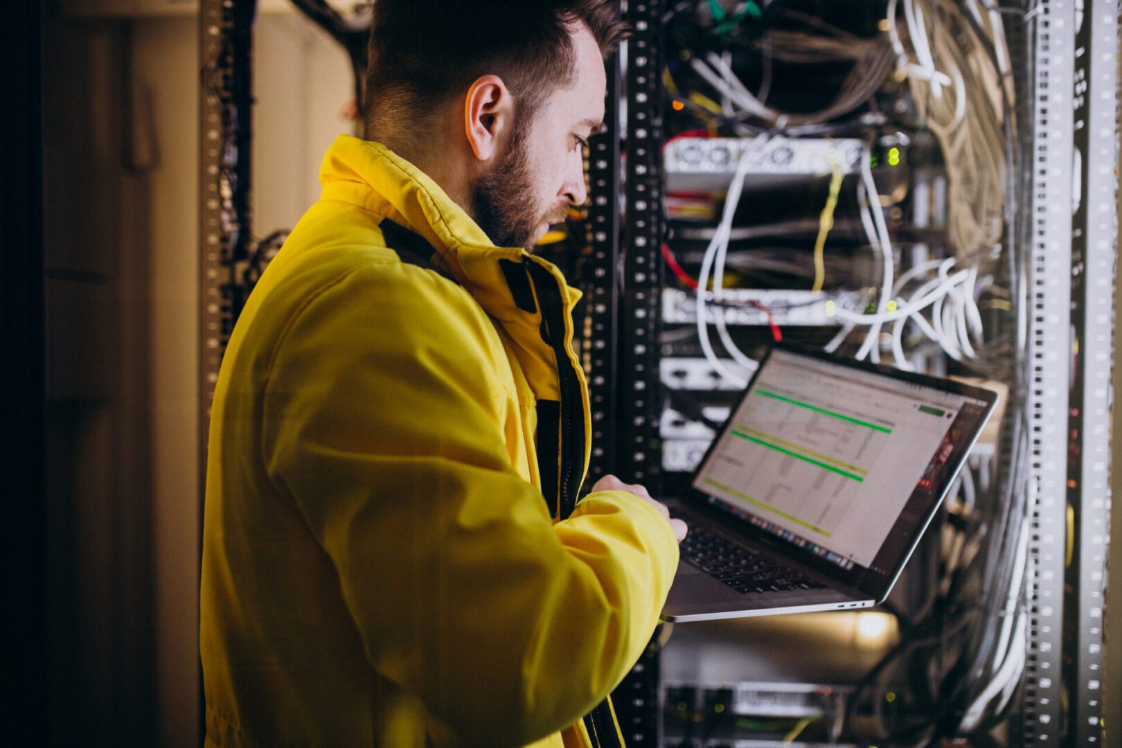 The image shows a man in a yellow jacket working in a server room or data center. He is holding a laptop and appears to be checking data or configurations, possibly performing system diagnostics or network maintenance. The background features a server rack filled with network cables and various hardware components, indicating a technical or IT environment. The screen of the laptop displays a spreadsheet, suggesting he might be referencing or updating logs or inventory data.