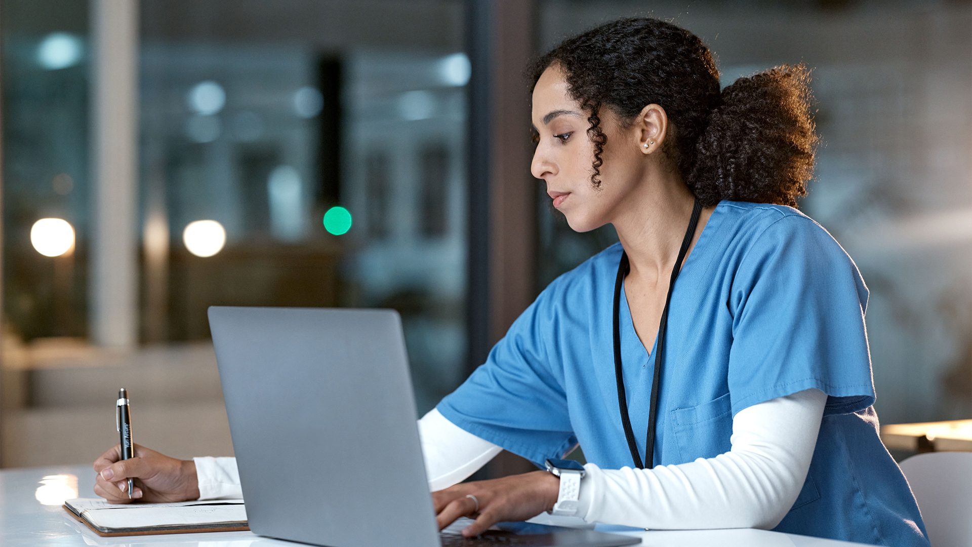 The image shows a healthcare professional, likely a nurse or medical assistant, working at a desk. She is wearing blue scrubs and a white undershirt, with a lanyard around her neck. She appears focused, writing notes on a notepad while looking at a laptop screen, possibly reviewing patient records or conducting research. The setting seems to be indoors, possibly in a hospital or clinic, and it looks like it might be evening based on the lighting and reflections in the background.