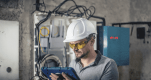 A male electrician or engineer wearing a white hard hat and yellow safety glasses is inspecting a blue tablet while working in an industrial setting. He has a focused expression as he examines data or schematics. Behind him, an open electrical panel with tangled wires and a yellow cable loop is visible, along with a blue electrical control box displaying red digital numbers. The setting has exposed concrete walls, giving it a rugged, workshop-like appearance.