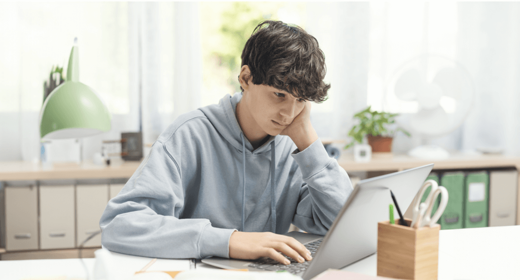 A young male student with dark curly hair and a light blue hoodie sits at a desk, looking at his laptop with a focused yet slightly tired expression. His hand rests on his cheek as he leans forward. The desk is organized with school supplies, including a notebook, pens, and a wooden container holding scissors. In the background, a bright study space with shelves, a green desk lamp, a potted plant, and a fan is visible. Natural light streams in through a window, creating a calm and studious atmosphere.