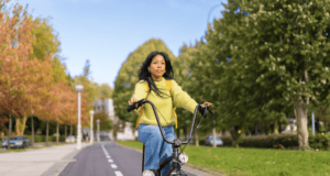 A young woman with long black hair rides a black bicycle down a dedicated bike lane in an urban park. She is wearing a fuzzy yellow sweater, blue jeans, and a yellow backpack. Her expression is calm and focused as she enjoys the ride. The background features lush green trees, some with autumn-colored leaves, a clear blue sky, lampposts, and parked cars along the street.