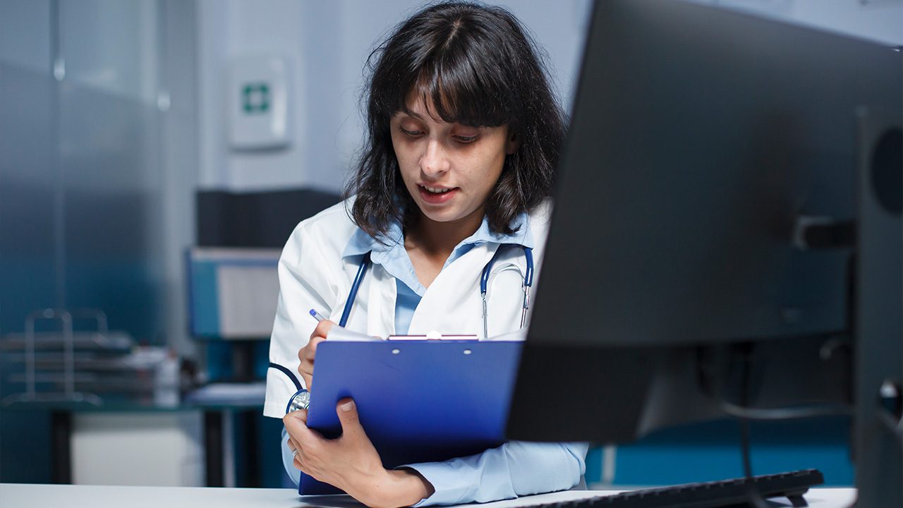 A female doctor with shoulder-length dark hair, wearing a white lab coat and a stethoscope around her neck, is sitting at a desk in a medical office. She holds a blue clipboard and pen, appearing focused as she reviews or takes notes. A large computer monitor partially obscures her view. The background features a clinical setting with office equipment and shelves, suggesting a professional healthcare environment.