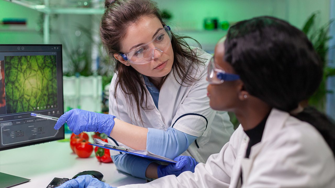 Two female scientists wearing protective eyewear, lab coats, and blue gloves are working in a laboratory. One scientist, holding a clipboard, is pointing at a computer screen displaying a magnified image of plant cells, while the other listens attentively. Fresh tomatoes are visible on the table, suggesting research related to food or agriculture. The background features green lighting and lab equipment, emphasizing a high-tech research environment.