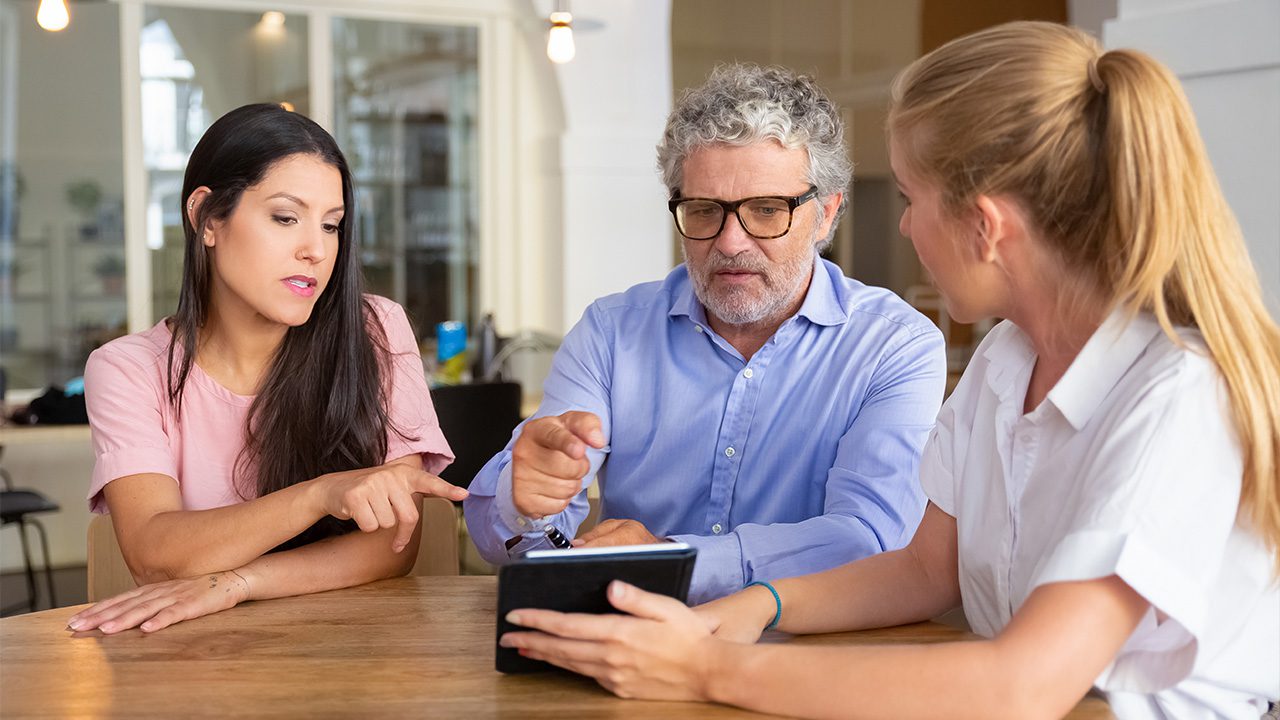 Three people, two women and one older man with gray hair and glasses, are sitting at a wooden table in a well-lit modern office or café. The man, wearing a light blue shirt, is gesturing towards a tablet held by the blonde woman in a white blouse. The brunette woman in a pink top is also pointing at the screen, appearing engaged in the discussion. The background features shelves, glass windows, and warm lighting, suggesting a collaborative or business meeting setting.