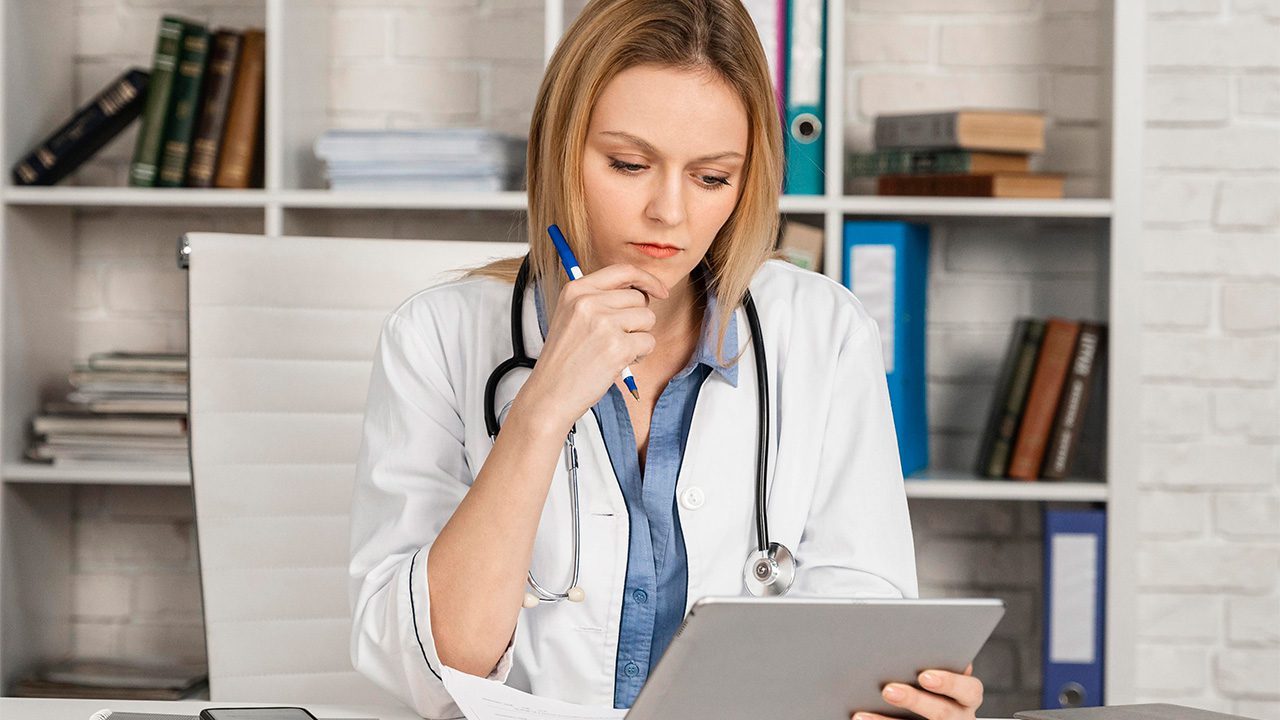 A female doctor with blonde hair, wearing a white lab coat and a stethoscope around her neck, sits at a desk in a medical office. She holds a pen to her chin while attentively reviewing information on a tablet. Behind her, a white bookshelf filled with medical books, binders, and stacks of documents is visible. The setting suggests a professional and focused work environment.