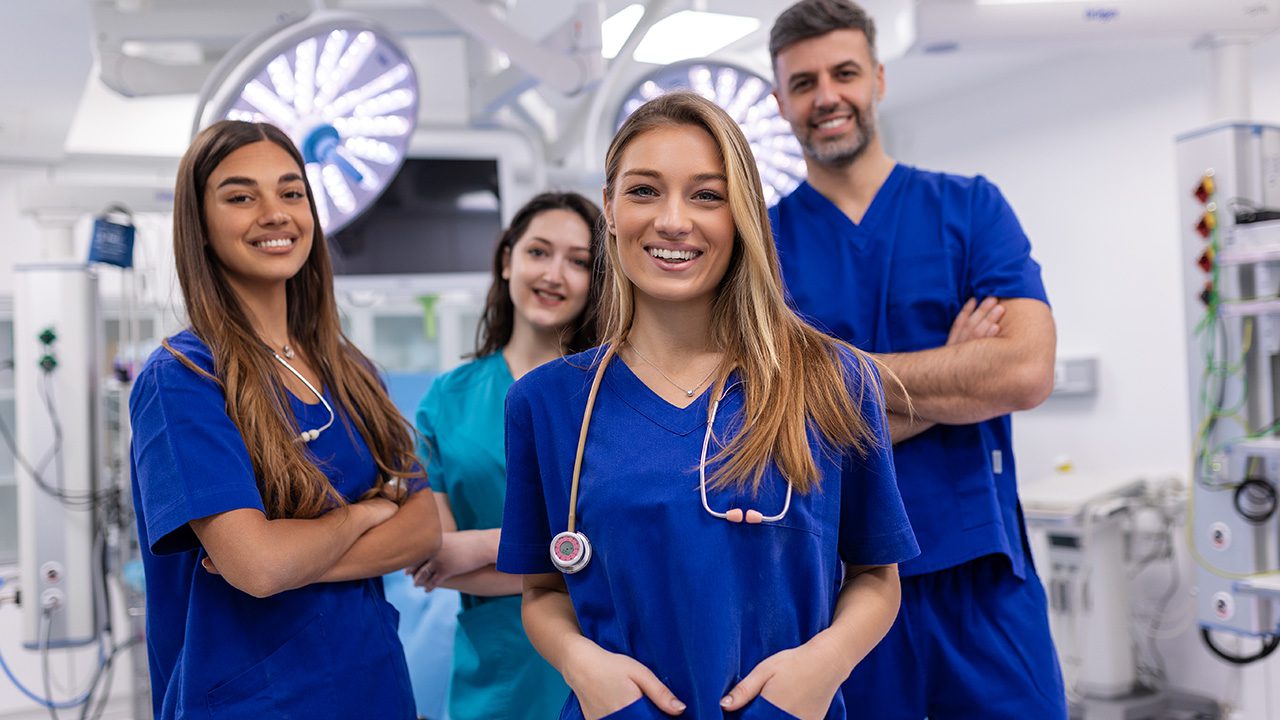 A group of four medical professionals, three women and one man, stand confidently in an operating room, dressed in blue scrubs. The woman in the foreground has long blonde hair, a stethoscope draped around her neck, and is smiling warmly at the camera. To her left, another woman with long brown hair stands with her arms crossed, also smiling. Behind them, a woman in teal scrubs and a man with short graying hair, his arms folded, are both smiling. The background features surgical lights, medical monitors, and various hospital equipment, indicating a clinical setting.