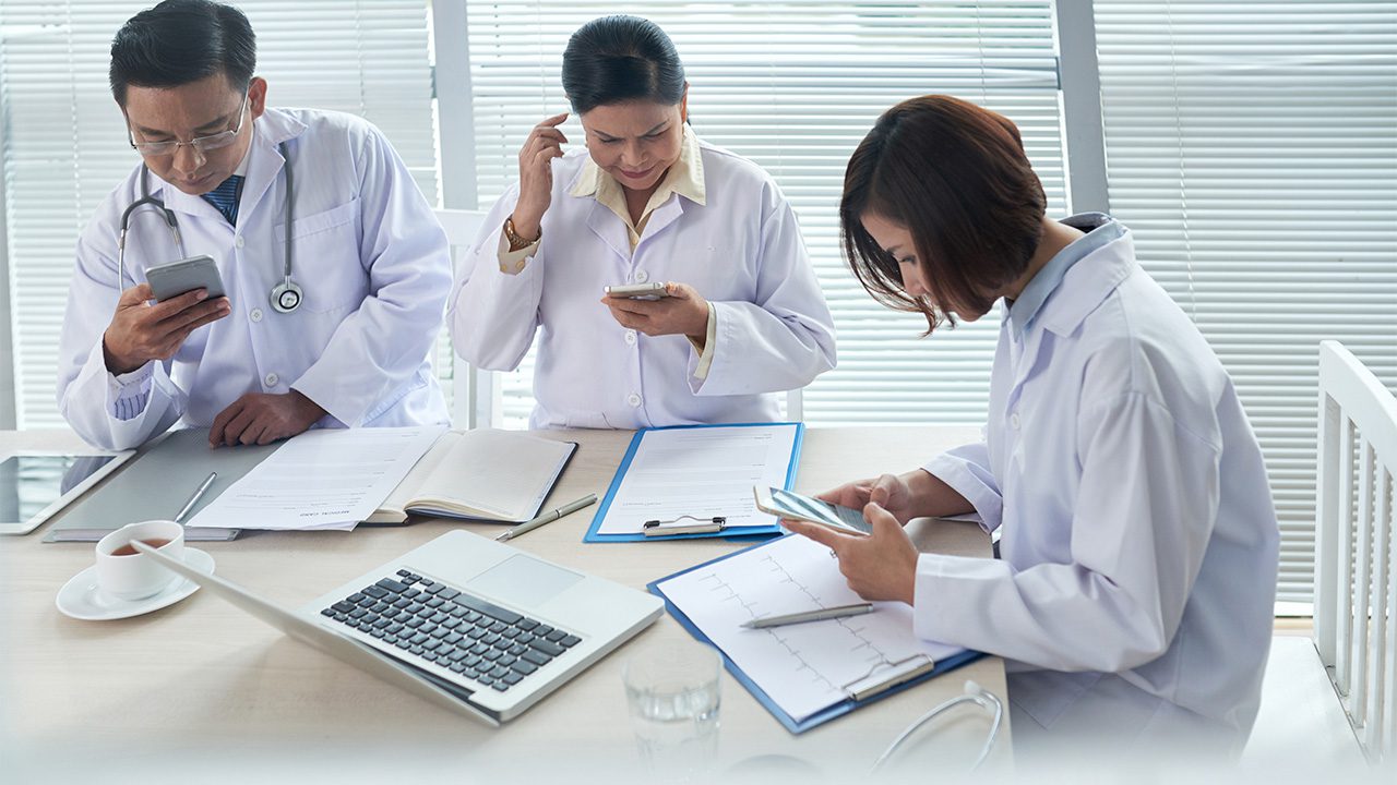 Three medical professionals in white lab coats are gathered around a table in a well-lit office with closed blinds. They are all focused on their smartphones. The male doctor on the left, wearing glasses and a stethoscope around his neck, is looking at his phone while holding a pen over some documents. The middle-aged female doctor in the center is also checking her phone while touching her ear. The younger female doctor on the right is reviewing medical charts on a clipboard while using her phone. The table is cluttered with open notebooks, a laptop, a pen, a cup of tea, and medical documents, including an ECG report.