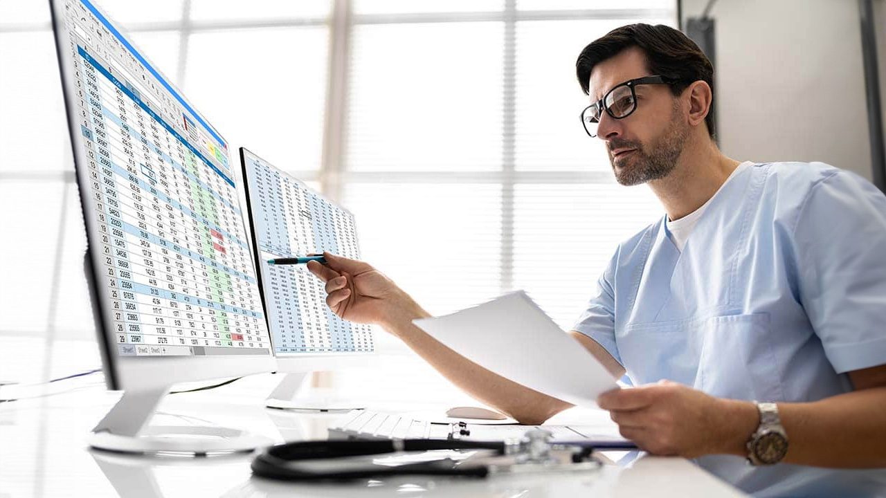 A male healthcare professional in light blue scrubs and glasses is analyzing data on two computer monitors displaying spreadsheets with medical or patient information. He holds a pen in one hand, pointing at the screen, and a document in the other. A stethoscope rests on the desk, indicating a medical setting. The bright office environment suggests a clinical or administrative workspace.