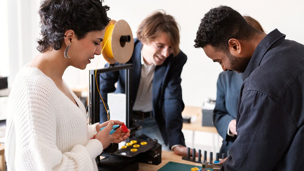 A group of four people collaborates in a workshop or laboratory setting, working with a 3D printer. A woman in a white sweater holds a small red and blue 3D-printed object, examining it closely. A man in a dark jacket leans forward, engaging in the project, while another man in a blazer smiles in the background. A fourth person, partially visible, observes the process. The workspace contains various tools, printed components, and a spool of filament feeding into the 3D printer.