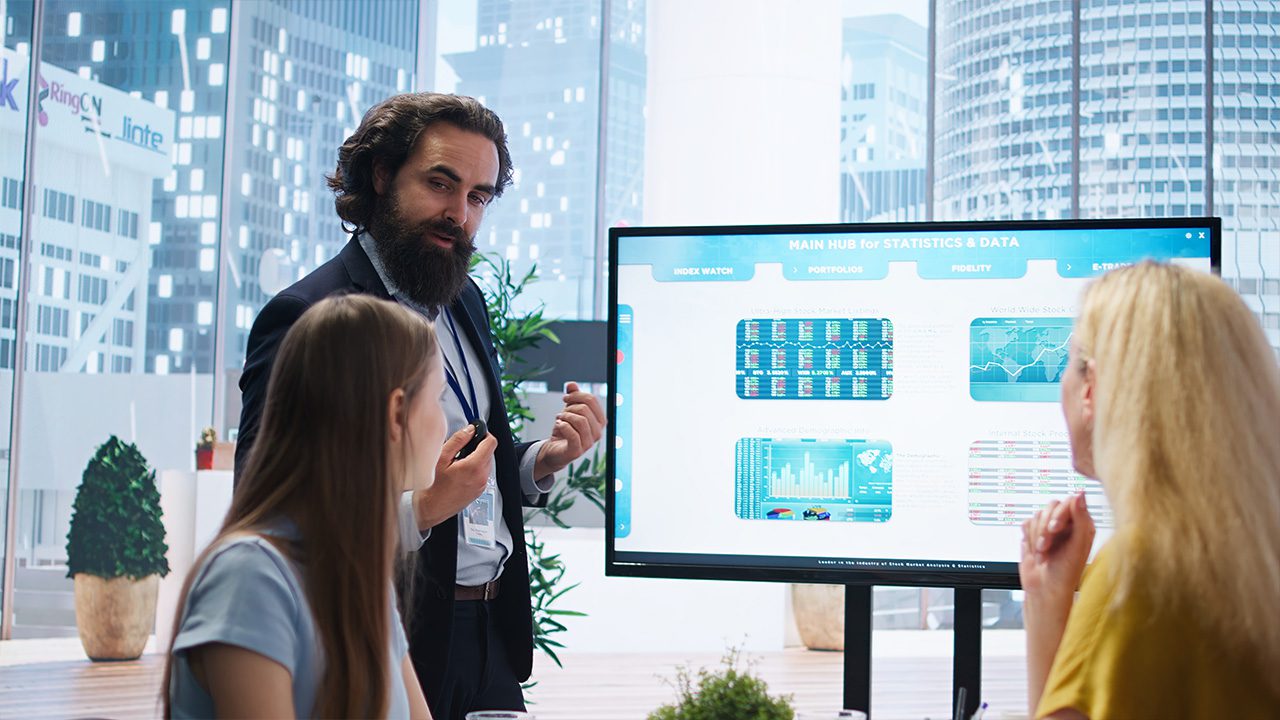 A bearded man in a suit and lanyard gives a presentation to two women in a modern office with large windows overlooking a cityscape. He gestures while explaining financial data displayed on a large screen, which includes charts, graphs, and stock market statistics. The two women, one with long brown hair and the other with blonde hair, listen attentively. The office features potted plants and contemporary furnishings.