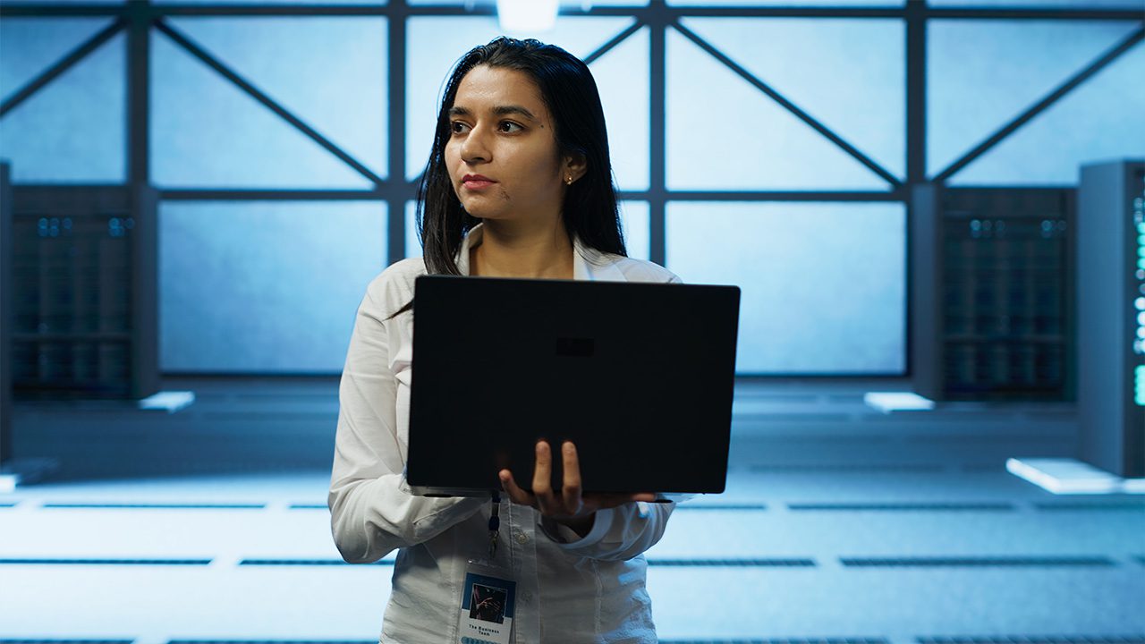 A woman with long dark hair, wearing a white shirt and a lanyard with an ID badge, stands in a high-tech server room holding a black laptop. She has a focused expression as she looks off to the side. The background features large illuminated panels and rows of server racks with blinking lights, creating a futuristic and data-driven atmosphere.