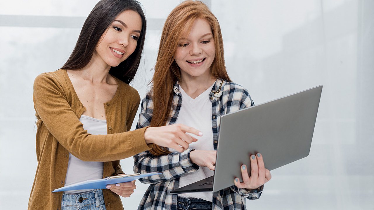 Two young women stand together, smiling and looking at a laptop screen. The woman on the left, wearing a brown cardigan over a white top, holds a clipboard and points at the screen. The woman on the right, wearing a plaid shirt over a white t-shirt, holds the laptop with one hand while typing with the other. They appear engaged and excited, possibly discussing a project or assignment. The background is bright and minimalistic, with large windows letting in natural light.