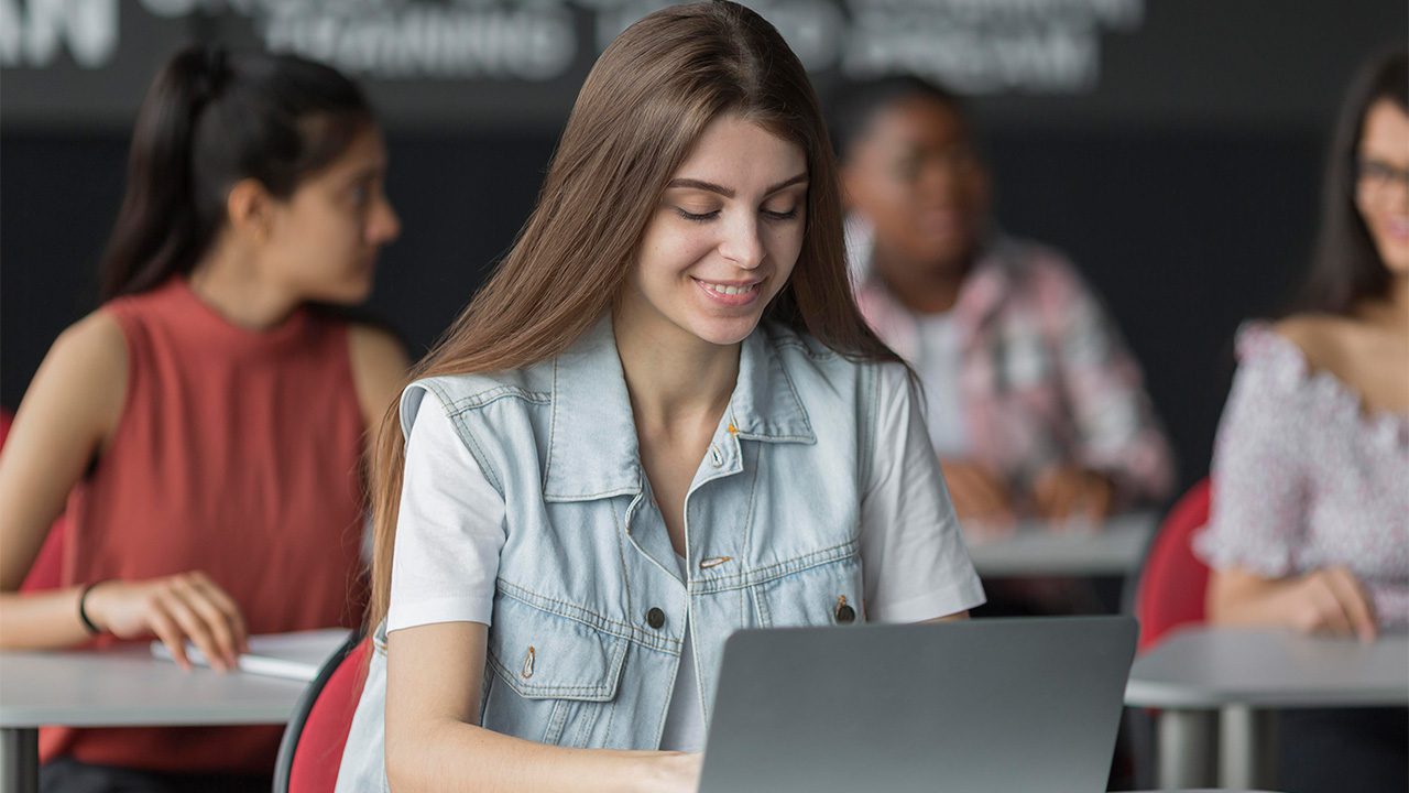 A young woman with long brown hair and light skin is sitting in a classroom, smiling while working on a laptop. She is wearing a white t-shirt with a light blue denim vest. Other students in the background are seated at desks, engaged in conversations or their own tasks. The setting appears to be a modern educational environment, possibly a university or training center, with a diverse group of students. The image conveys a sense of focus, learning, and engagement in a collaborative classroom setting.