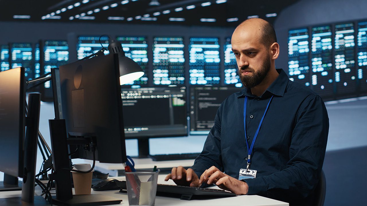 A bald man with a beard, wearing a dark blue shirt and a lanyard with an ID badge, is working on a computer in a high-tech security or IT environment. He has a serious and focused expression as he types on a keyboard. Multiple monitors display lines of code or system data, and a dim desk lamp provides additional lighting. The background features a large digital screen or server room with rows of illuminated data panels, suggesting a cybersecurity, IT operations, or network administration setting. A coffee cup and office supplies are on the desk, indicating a long work session.