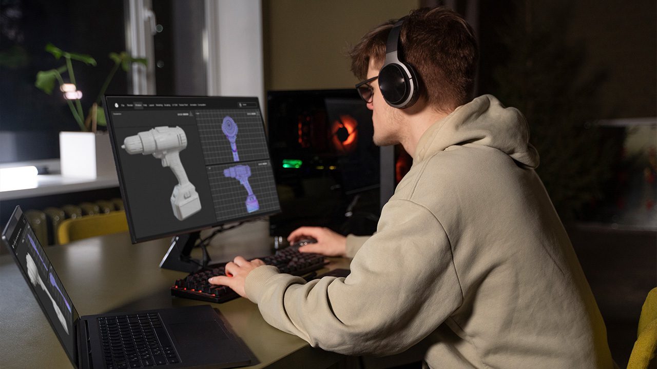 A young man wearing a beige hoodie, glasses, and large over-ear headphones is working on a 3D modeling project at his computer. He is seated at a desk with a dual-monitor setup, using a keyboard and mouse to design a detailed 3D model of a power drill displayed on the screen. A laptop beside him shows a similar 3D rendering. The dimly lit room has a modern setup with a gaming-style desktop PC glowing in the background, and a plant near a window adds a touch of greenery. The scene suggests a digital artist or engineer engaged in 3D design or game development.