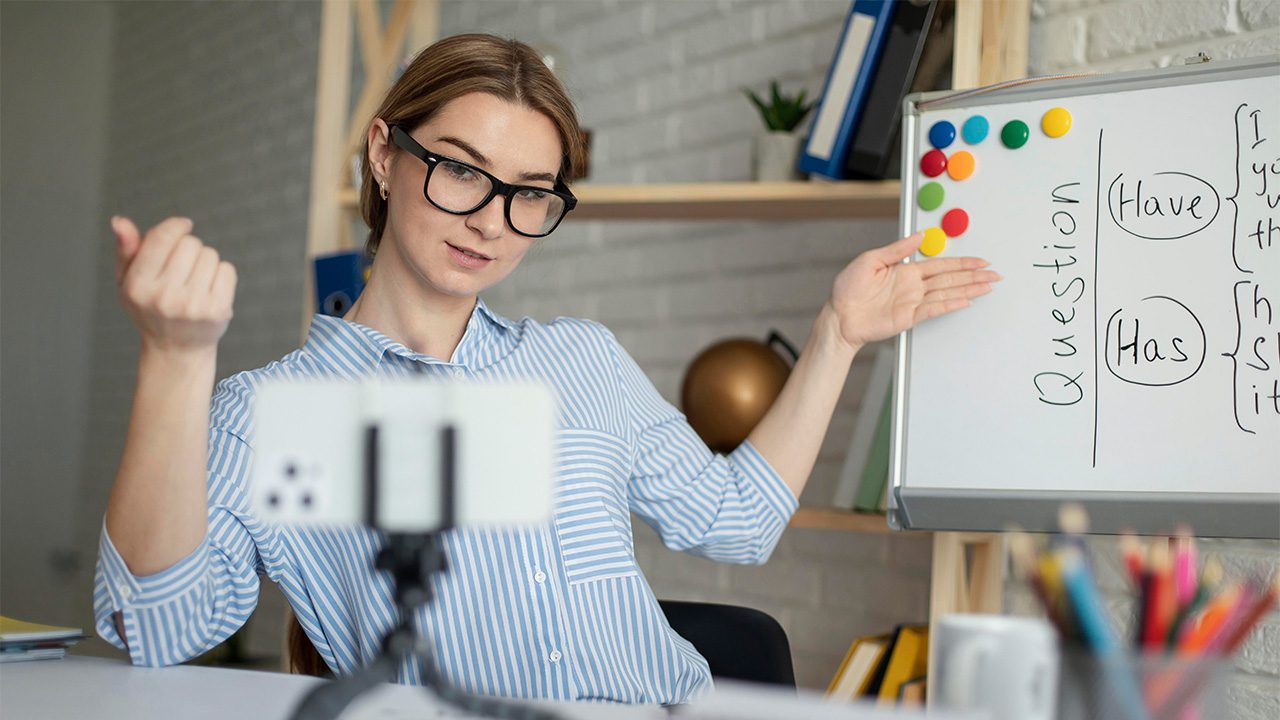 A woman with light skin, glasses, and brown hair tied back is conducting an online lesson, gesturing towards a whiteboard while speaking to a smartphone mounted on a tripod. She is wearing a blue and white striped shirt and has an engaged expression. The whiteboard behind her contains a grammar lesson, showing the words "Have" and "Has" under the heading "Question," with colorful magnets attached. The background features a neatly organized home office or classroom setting with books, folders, and a decorative globe on shelves. The foreground includes a desk with pencils and stationery, indicating a teaching environment.