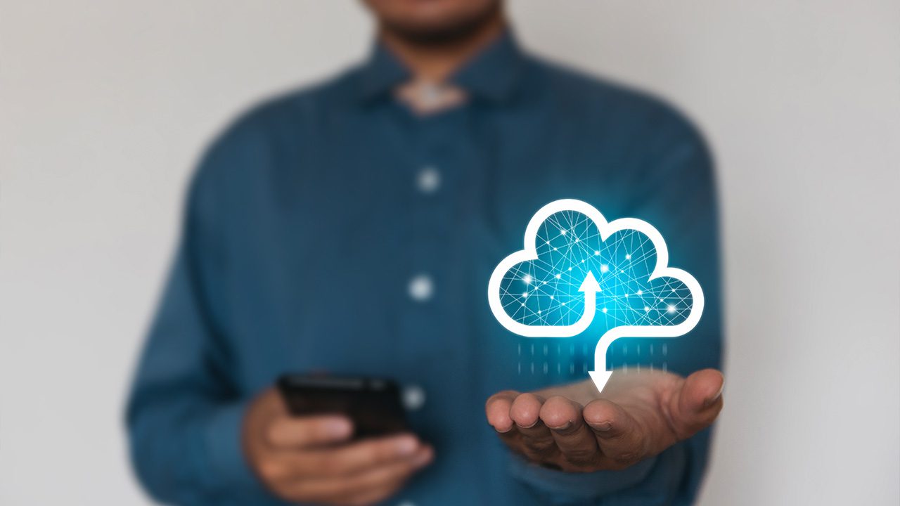 A person wearing a blue shirt holds out their hand with a glowing digital icon of a cloud hovering above it. The cloud features interconnected lines and nodes, symbolizing network connectivity, with an upward arrow in its center representing cloud computing or data upload. The individual holds a smartphone in their other hand, partially blurred, emphasizing a focus on technology and digital innovation. The background is plain and neutral.