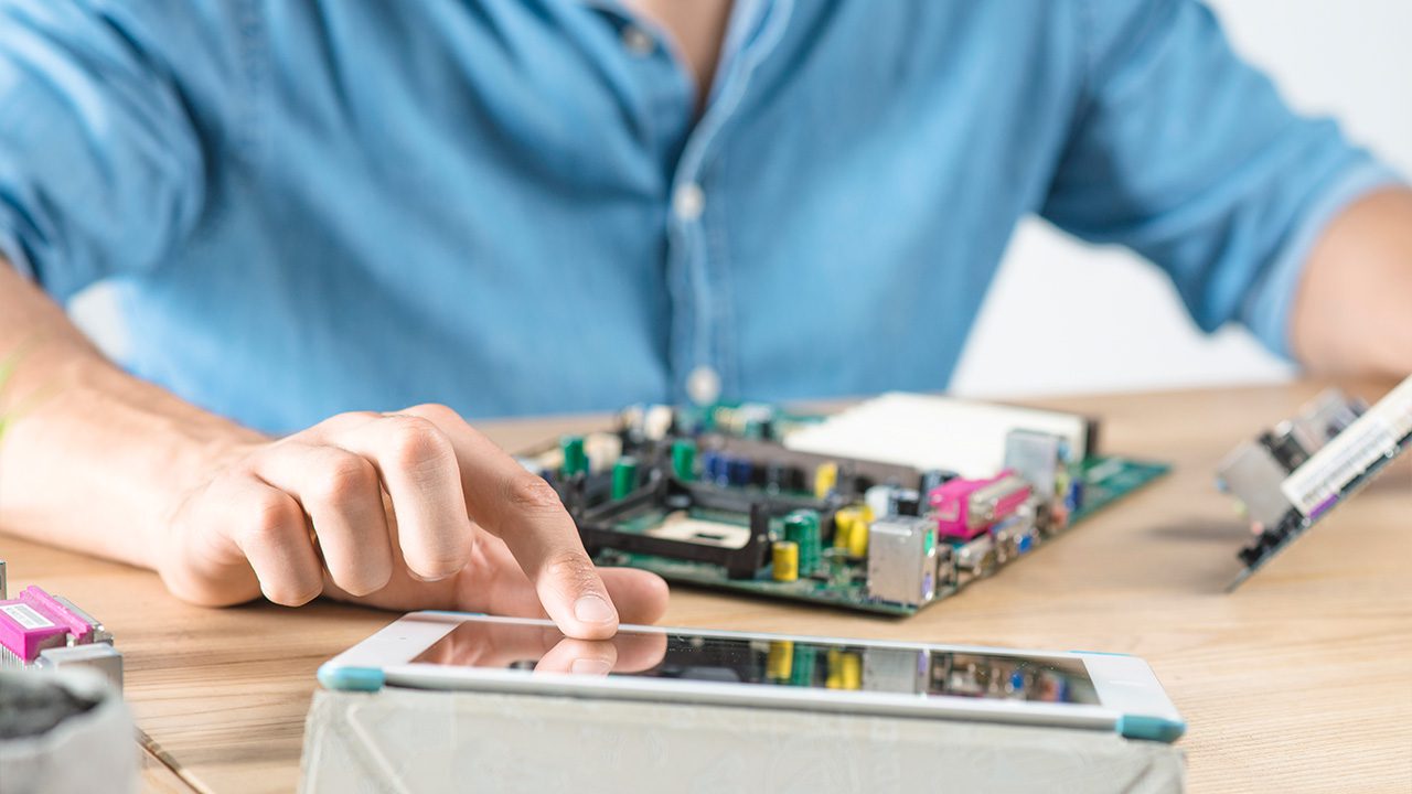 A person wearing a blue shirt is working on assembling or repairing a computer motherboard at a wooden table. Their left hand holds a computer component, while their right hand interacts with a tablet placed on a stand. The table is scattered with various electronic parts, including a motherboard and other circuit components, suggesting a tech repair or hardware development setting.