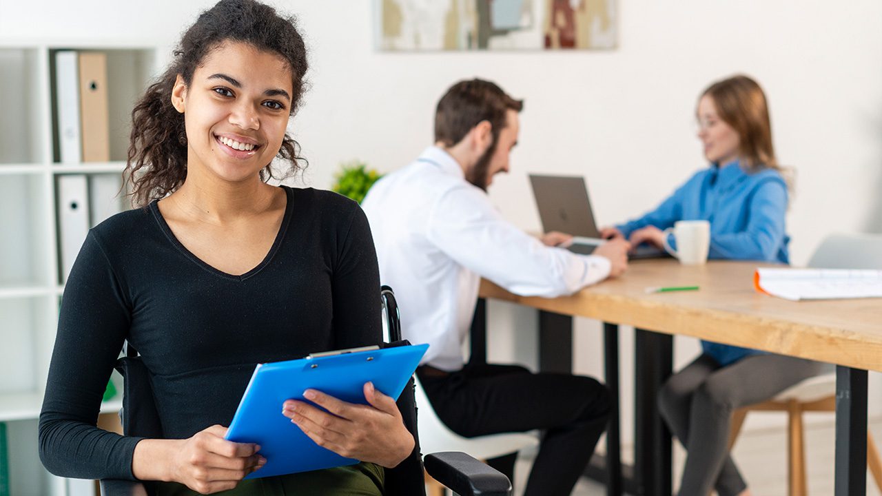 A young woman with curly hair, wearing a black top, sits in a wheelchair holding a blue clipboard and smiling at the camera. In the background, a man in a white shirt and a woman in a blue blouse are seated at a wooden table, working on laptops. The office space has white walls, shelves with binders, and a minimalist decor. The setting conveys a professional and inclusive work environment.