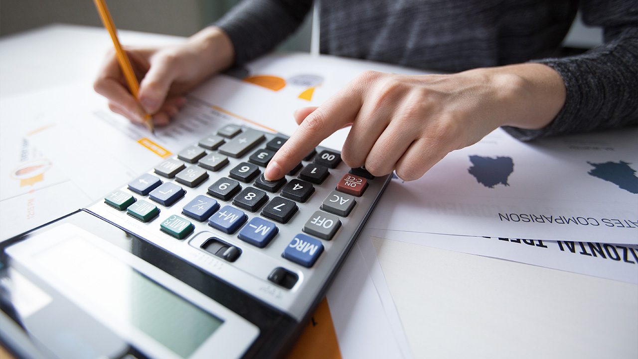 A close-up of a person's hands working with a calculator and financial documents. One hand presses a button on a large calculator, while the other holds a pencil and writes on a sheet of paper with charts and graphs. The documents contain text and graphics related to financial analysis or data comparison. The scene suggests financial planning, budgeting, or accounting work.