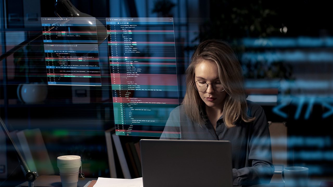 A young woman with glasses and blonde hair sits at a desk, focused on her laptop screen in a dimly lit workspace. Transparent digital code overlays float in front of her, displaying lines of programming in a futuristic style. The background includes a desk lamp, bookshelves, and a coffee cup, suggesting a late-night coding or cybersecurity session.