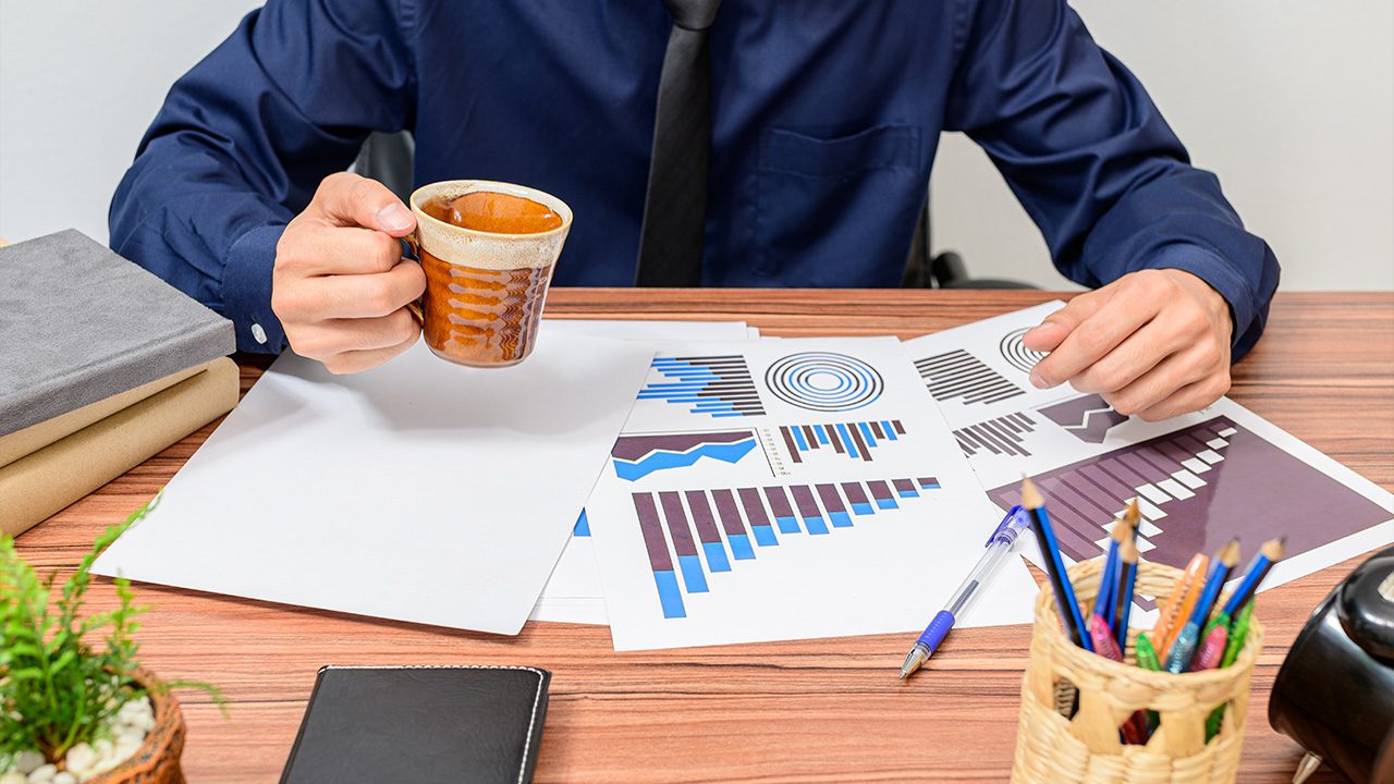 A businessman wearing a dark blue shirt and black tie sits at a wooden desk, holding a cup of coffee while reviewing printed charts and graphs with blue and brown data visualizations. The desk is neatly organized with a pen, a notebook, a stack of books, a small potted plant, and a woven pencil holder filled with colorful pens and pencils. The setting suggests a professional work environment focused on data analysis or business strategy.