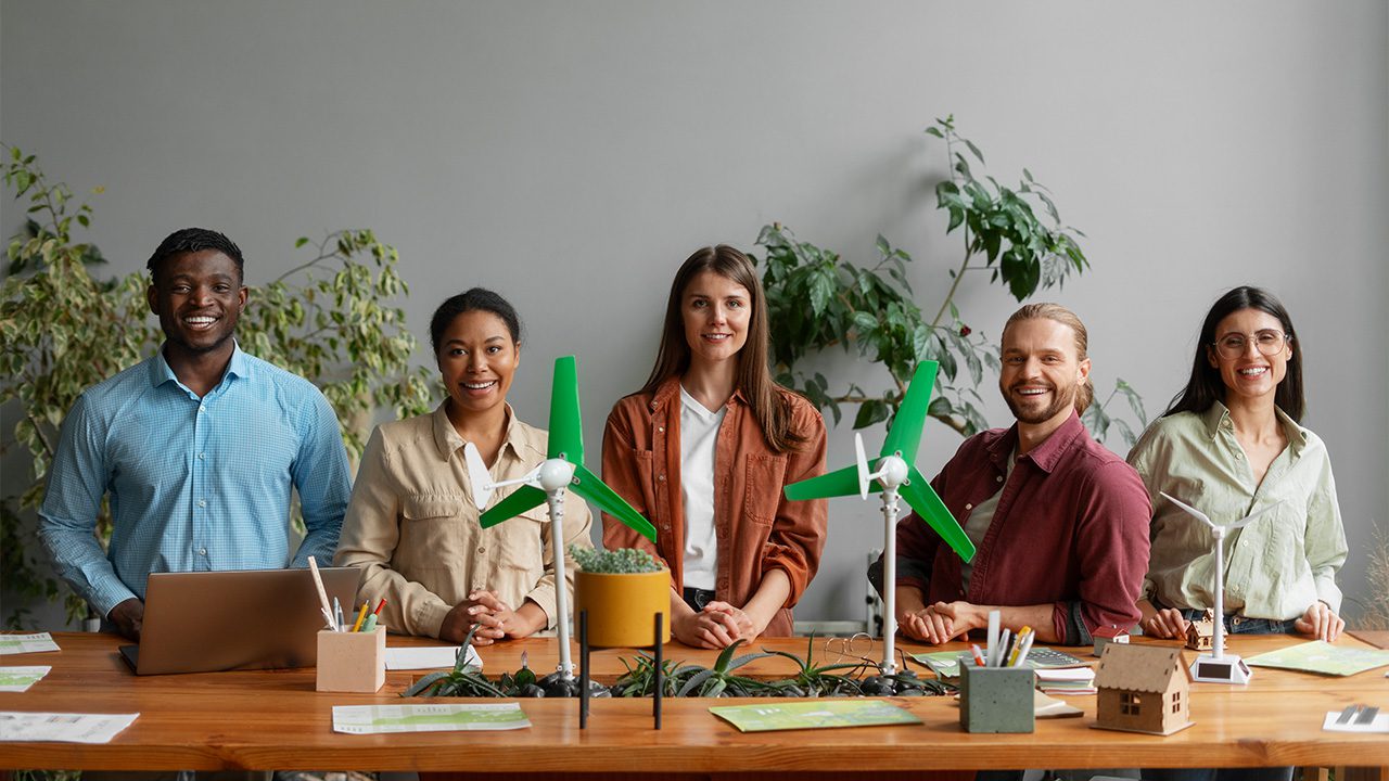 A diverse group of five professionals, three women and two men, stand behind a wooden table in a modern, eco-friendly workspace. They are smiling and looking at the camera. The table is decorated with small wind turbine models, green plants, documents, and office supplies, suggesting a focus on renewable energy or sustainability. One man on the left is working on a laptop, while the others stand confidently. The background features a minimalist interior with large green plants, enhancing the natural and environmental theme.