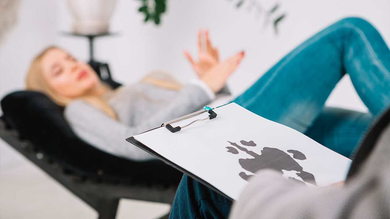 A woman with long blonde hair, wearing a gray sweater and blue jeans, reclines on a black chaise lounge in a therapy session. She gestures with her hands while speaking. In the foreground, a therapist, whose face is not visible, holds a clipboard with a Rorschach inkblot test. The setting is a modern, well-lit office with a neutral color scheme, suggesting a psychological consultation or counseling session.