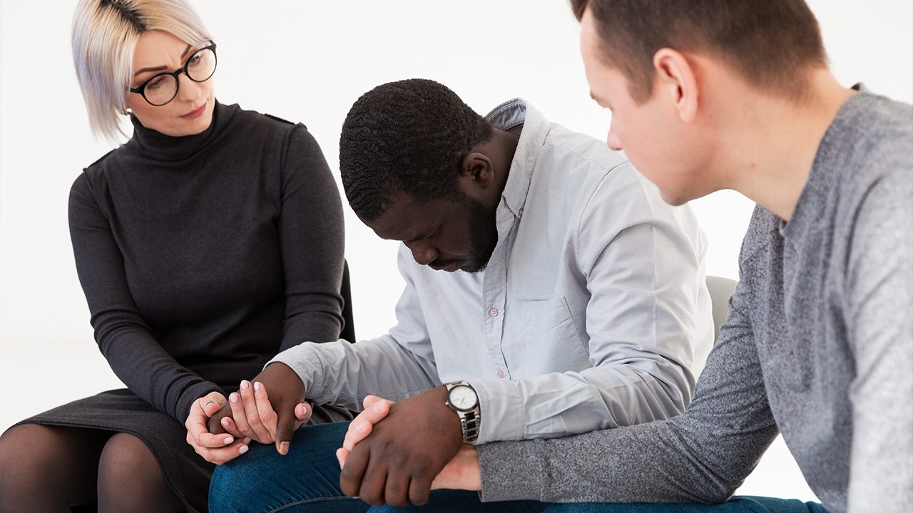 A distressed man in a light gray shirt sits with his head bowed, holding hands with two supportive individuals beside him. To his left, a woman with short blonde hair, glasses, and a black turtleneck dress gently holds his hand with a concerned expression. To his right, a man in a gray sweater leans in, offering comfort. The setting is a bright, minimalistic space, suggesting a support group or counseling session. The image conveys a sense of empathy, emotional support, and community.