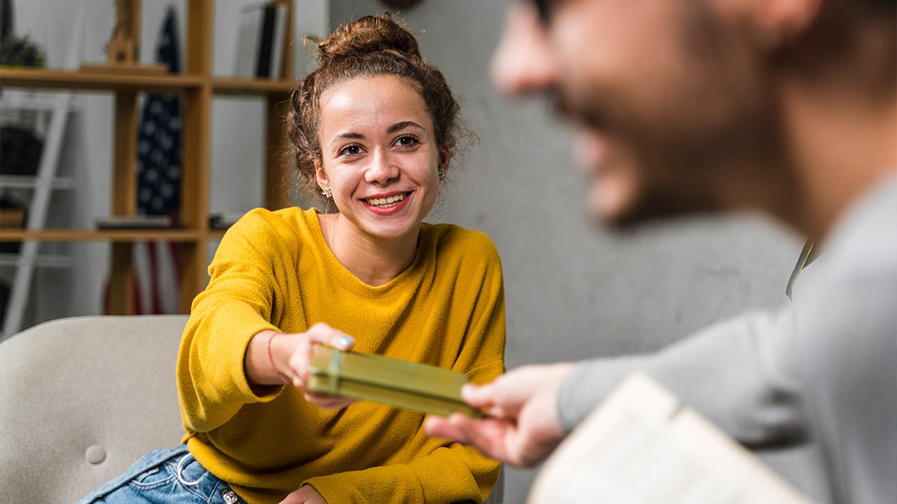 A young woman with curly hair, wearing a mustard-yellow sweater, smiles warmly as she hands a book to a man sitting across from her. The man, partially visible in the foreground, wears glasses and holds another book in his hands. The background features a cozy indoor setting with shelves, books, and an American flag, suggesting a relaxed learning or discussion environment.