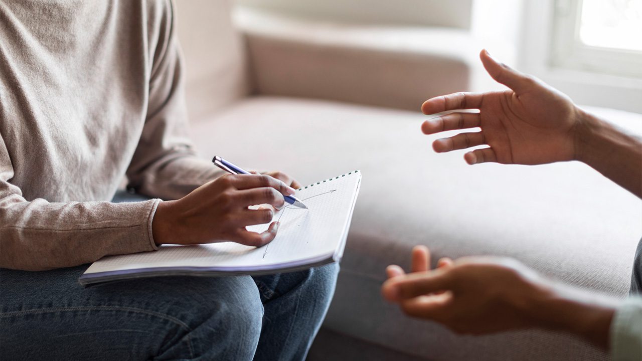 A close-up of a therapy or counseling session shows a therapist wearing a beige sweater, holding a pen and taking notes in a spiral notebook. The client, whose face is not visible, gestures with their hands while speaking. The setting is a comfortable room with a soft couch and natural lighting, suggesting a supportive and professional environment for discussion.