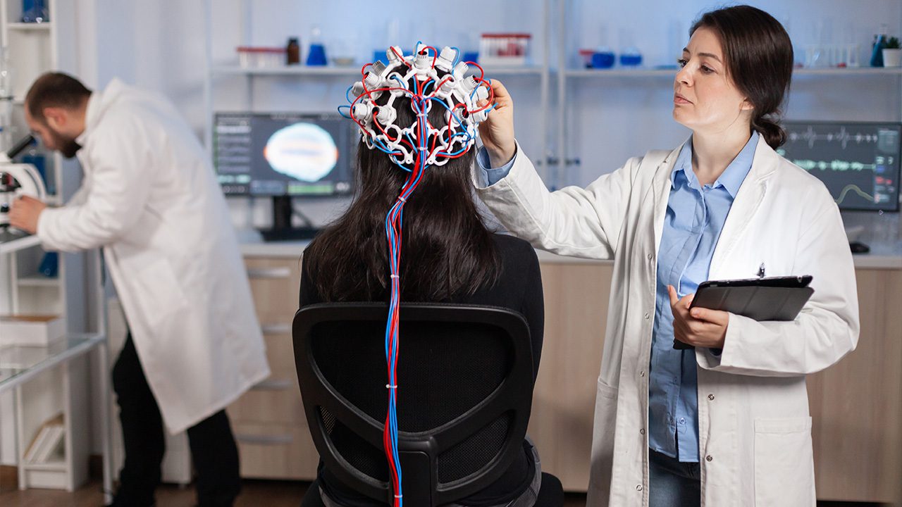 A female scientist in a white lab coat is conducting a brain study on a seated participant wearing an EEG cap with multiple electrodes and red and blue wires. She holds a tablet and adjusts the cap while monitoring the subject. In the background, another researcher in a lab coat is working with a microscope, and computer screens display brain scan data. The setting appears to be a high-tech neuroscience laboratory.