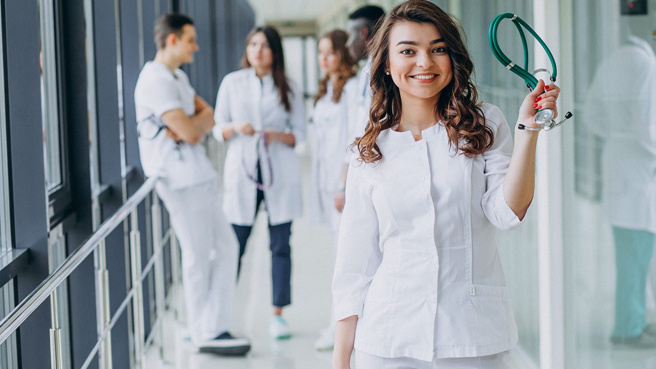 A young female medical professional in a white coat smiles while holding up a green stethoscope. She stands confidently in the foreground of a bright hospital corridor, with a group of fellow medical students or healthcare professionals engaged in conversation in the background.