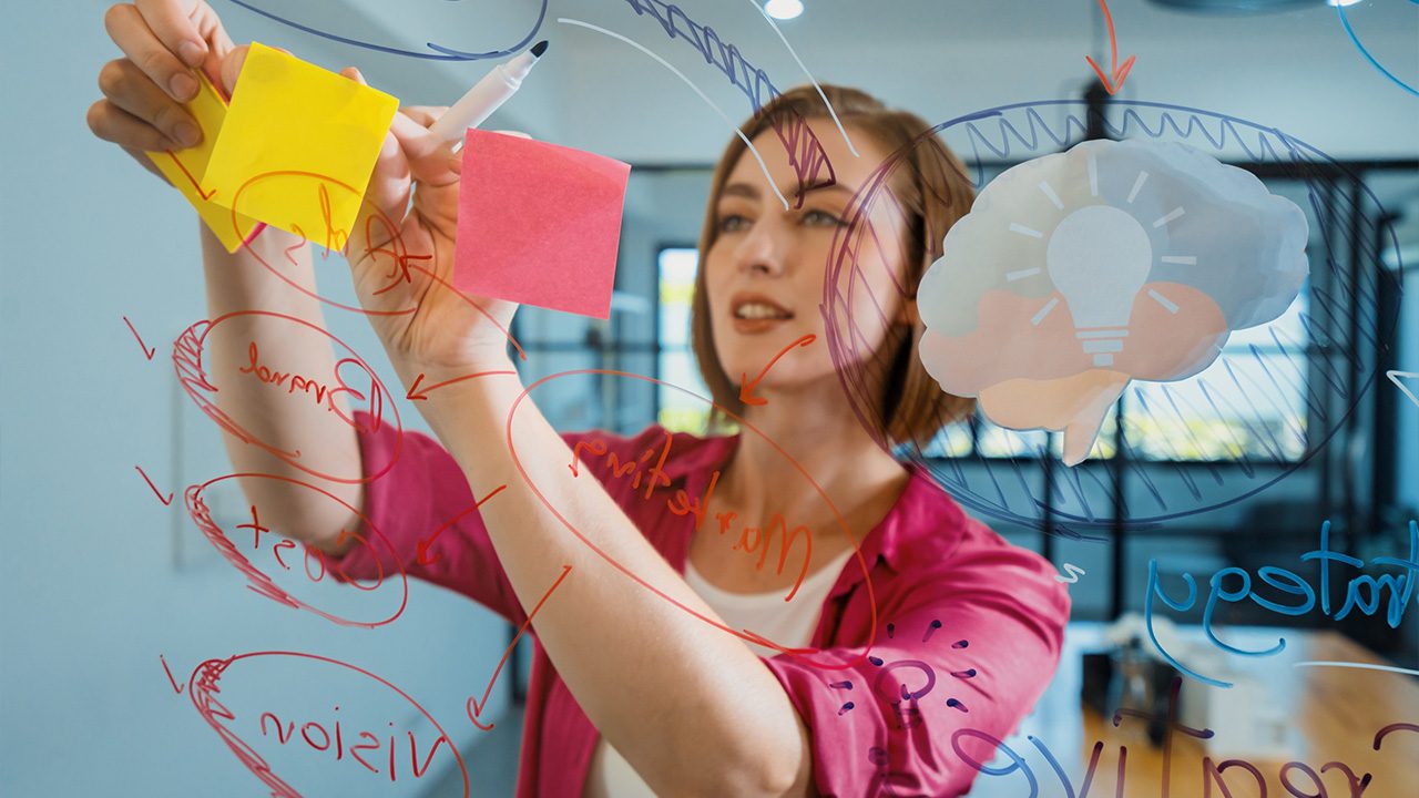 A woman in a pink blouse is brainstorming ideas on a transparent glass board, writing with a marker and attaching colorful sticky notes. The board is filled with handwritten words, diagrams, and a drawing of a brain with a lightbulb inside, symbolizing creativity and innovation. The modern office setting in the background suggests a collaborative and dynamic work environment.